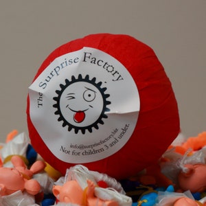 May include: A red paper ball with a white label that says "The Surprise Factory" and has a cartoon face with a wink and tongue sticking out. The label also says "info@surprisefactory.biz Not for children 3 and under."