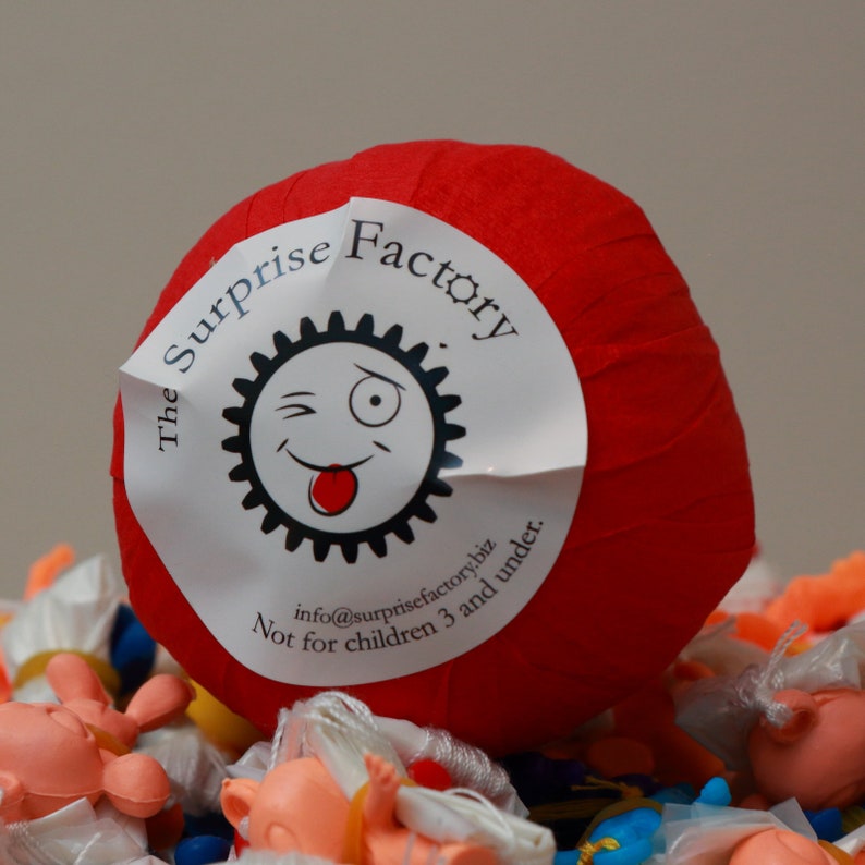 May include: A red paper ball with a white label that says "The Surprise Factory" and has a cartoon face with a wink and tongue sticking out. The label also says "info@surprisefactory.biz Not for children 3 and under."