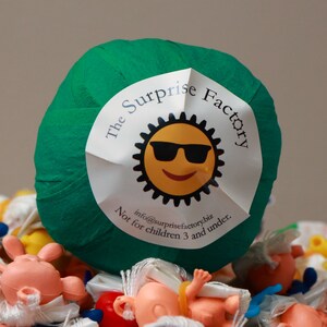 May include: A green ball wrapped in tape with a white label that says "The Surprise Factory" with a cartoon sun wearing sunglasses and the text "info@surprisefactory.biz Not for children 3 and under."