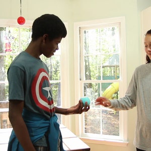 May include: A young person wearing a blue shirt with a red, white, and blue star design is handing a blue and green ball to another young person wearing a gray long-sleeved shirt.
