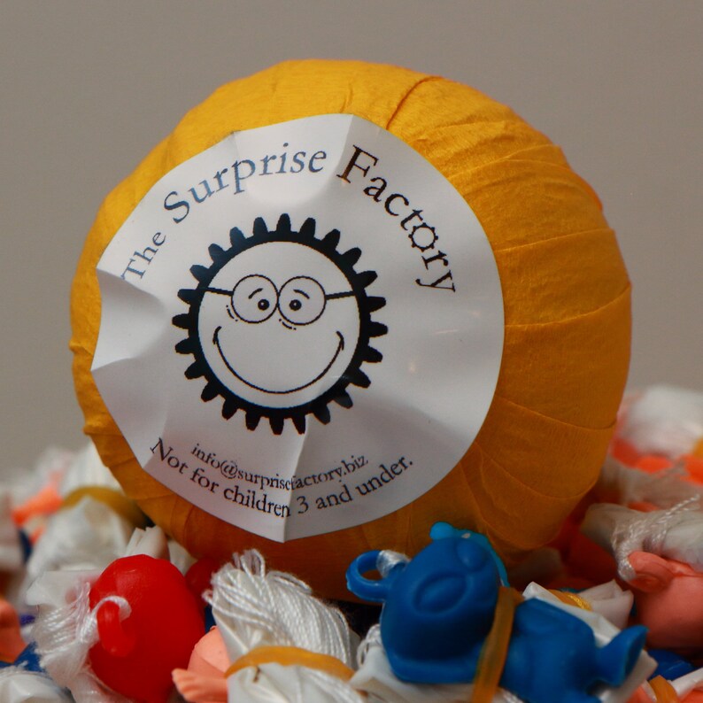 May include: A yellow ball with a white label that says "The Surprise Factory" and a cartoon sun with a smiling face. The label also says "Not for children 3 and under." and "info@surprisefactory.biz".