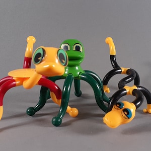 May include: Three colorful bendable toys shaped like animals. The first toy is yellow and red with green eyes and a long snout. The second toy is green with green eyes and a wide mouth. The third toy is black and yellow with blue eyes and a long snout.
