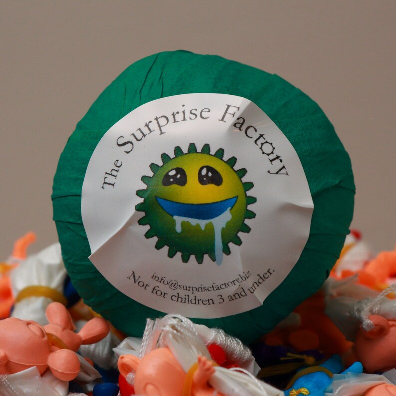 May include: A green ball with a white label that says "The Surprise Factory" and features a cartoon green, yellow, and blue smiley face. The text on the label also says "info@surprisefactory.biz. Not for children 3 and under."