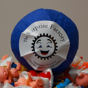 May include: A blue wrapped ball with a white label that says "The Surprise Factory" with a smiling sun logo and the text "info@surprisefactory.biz Not for children 3 and under."