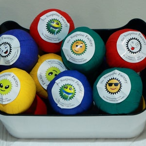 May include: A white basket filled with colorful fabric balls. Each ball has a white label with a different smiley face and the text "The Surprise Factory".