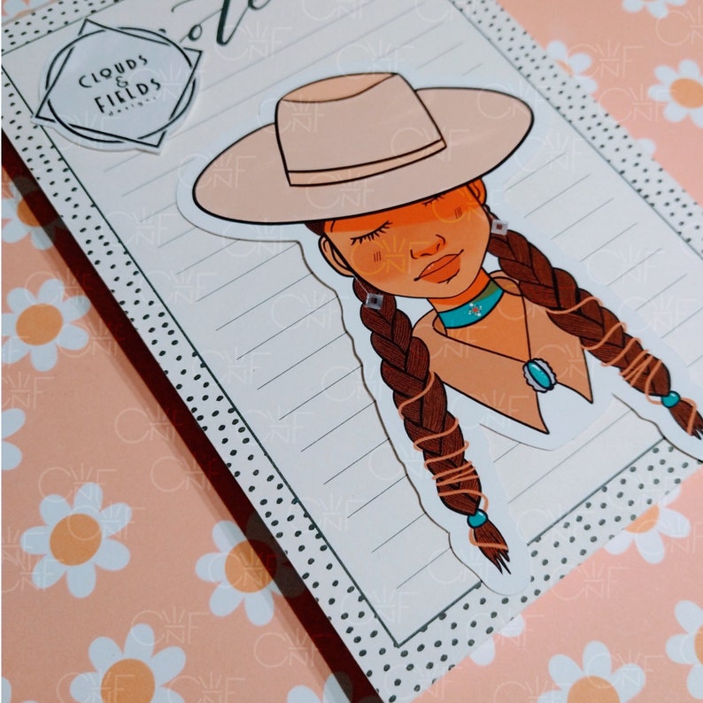 May include: A sticker of a cartoon girl with brown hair in braids, wearing a tan cowboy hat and a brown shirt with a turquoise necklace. The sticker is on a lined sheet of paper with a black and white polka dot border.