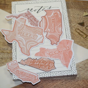 May include: A set of six pink and white state outline stickers with a doodle pattern. The stickers include California, Texas, Florida, Illinois, Kentucky, and New York.