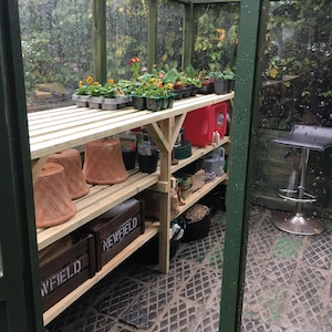 May include: A wooden greenhouse with shelves filled with potted plants, gardening supplies, and a wooden crate labelled "Newfield". The floor is covered in a diamond-patterned mat.