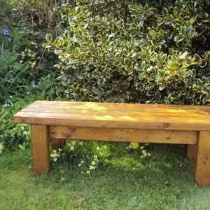 May include: A wooden bench with a natural brown finish sits on a grassy lawn in front of a green bush with yellow leaves.