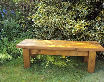 May include: A wooden bench with a natural brown finish sits on a grassy lawn in front of a green bush with yellow leaves.