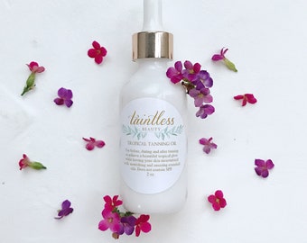 May include: A white bottle of "Tropical Tanning Oil" with a gold cap and dropper, surrounded by small pink and purple flowers. The label reads "taintless BEAUTY" and includes product information. The bottle contains 59 ml of product.