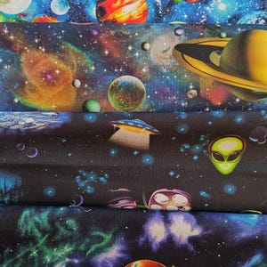 May include: Fabric with a vibrant space-themed design. The fabric features planets, galaxies, and stars in shades of blue, orange, and purple. The design includes a UFO and an alien.
