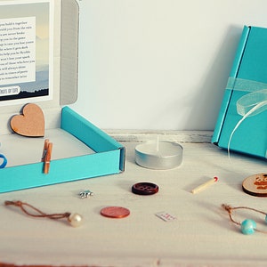 May include: A teal blue box with a white ribbon and a poem inside. The box is open and contains a wooden heart, a blue rubber band, a wooden clothespin, a small silver charm, a pearl, a copper coin, a brown button, a white tea light candle, a matchstick, a wooden bead necklace, and a wooden circle with a bear design.