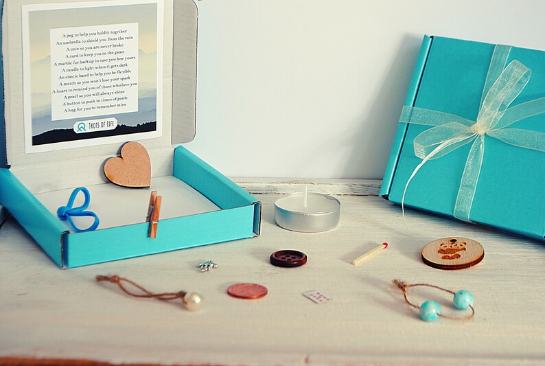 May include: A teal blue box with a white ribbon and a poem inside. The box is open and contains a wooden heart, a blue rubber band, a wooden clothespin, a small silver charm, a pearl, a copper coin, a brown button, a white tea light candle, a matchstick, a wooden bead necklace, and a wooden circle with a bear design.