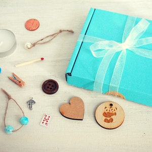 May include: A teal blue gift box with a white ribbon and a white bow. The box is open and contains a variety of small items, including a wooden heart, a wooden panda charm, a small wooden button, a teal blue string of beads, a small silver charm, a matchstick, a small wooden clothespin, a teal blue ribbon, a small candle, and a small wooden coin.