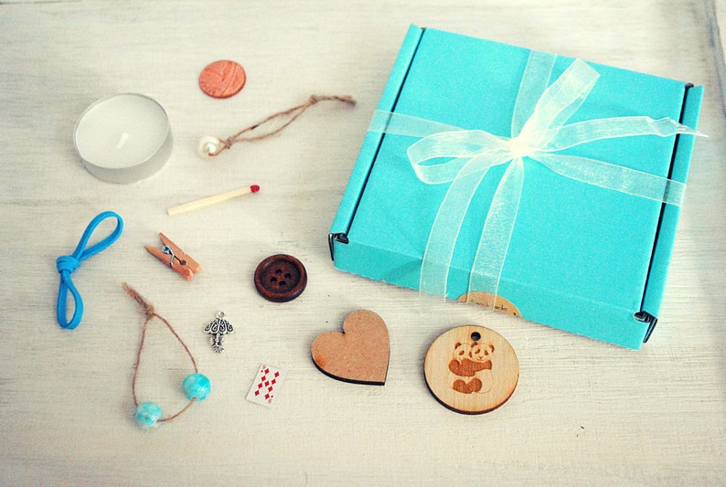 May include: A teal blue gift box with a white ribbon and a white bow. The box is open and contains a variety of small items, including a wooden heart, a wooden panda charm, a small wooden button, a teal blue string of beads, a small silver charm, a matchstick, a small wooden clothespin, a teal blue ribbon, a small candle, and a small wooden coin.