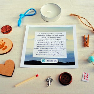 May include: A small white candle in a glass jar, a wooden heart, a panda bear charm, a blue rubber band, a coin, a match, a silver charm, a brown button, a playing card, a wooden clothespin, a pearl on a string, two blue beads on a string, and a card with a poem about the meaning of each item. The card reads "A peg to help you hold it together, An umbrella to shield you from the rain, A coin so you are never broke, A card to keep you in the game, A marble for luck in case you lose yours, A candle to light when it gets dark, An elastic band to help you be flexible, A match so you won't lose your spark, A heart to remind you of those who love you, A pearl so you will always shine, A button to push in times of panic, A hug to help you through hard times, TROTS OF LIFE".