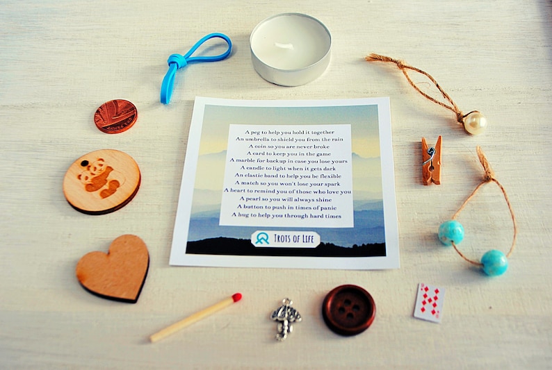 May include: A small white candle in a glass jar, a wooden heart, a panda bear charm, a blue rubber band, a coin, a match, a silver charm, a brown button, a playing card, a wooden clothespin, a pearl on a string, two blue beads on a string, and a card with a poem about the meaning of each item. The card reads "A peg to help you hold it together, An umbrella to shield you from the rain, A coin so you are never broke, A card to keep you in the game, A marble for luck in case you lose yours, A candle to light when it gets dark, An elastic band to help you be flexible, A match so you won't lose your spark, A heart to remind you of those who love you, A pearl so you will always shine, A button to push in times of panic, A hug to help you through hard times, TROTS OF LIFE".