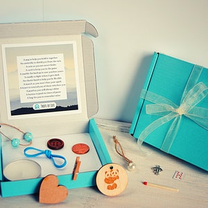 May include: A teal blue box with a white ribbon and a small card with a poem about tools for life. The box is open and contains a variety of small items, including a candle, a wooden heart, a wooden panda, a wooden button, a blue rubber band, a wooden clothespin, a small wooden bead necklace, a small white bead, a silver charm, a match, and a small deck of cards.
