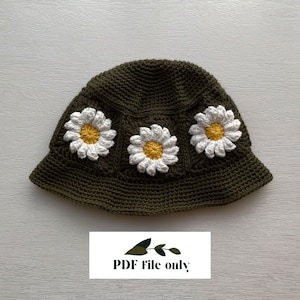 Puede incluir: Un sombrero de pescador de ganchillo verde oscuro adornado con tres aplicaciones de margaritas blancas y amarillas. El sombrero tiene un ala pequeña y se muestra sobre una superficie clara. El texto "PDF file only" es visible en la parte inferior.