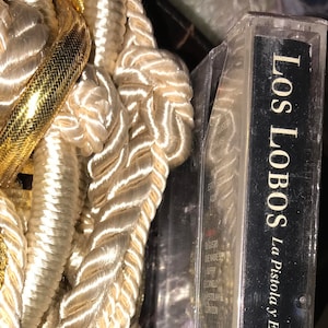 May include: A cassette tape with a black case and a white label. The label says "Los Lobos" and "La Pistola Y El Corazon". The tape is surrounded by white rope and gold trim.