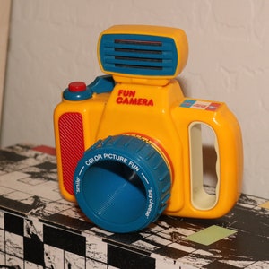 Peut inclure: Un appareil photo jouet jaune avec un objectif bleu et un bouton rouge. L'appareil photo a un flash bleu et blanc et indique "Fun Camera" sur le devant.