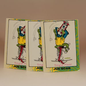 May include: Three trading cards featuring a cartoon character skateboarding. The character is green with a yellow hat and is wearing blue pants. The cards are white with green, yellow, and blue accents. The text on the cards reads "Tongue Plant!" and "Joe Scab".