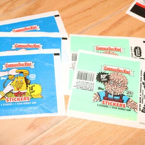 May include: A stack of Garbage Pail Kids sticker packs. The top pack features a blue background with a cartoon character holding a phone. The text on the pack reads "Garbage Pail Kids 14th Series Stickers 3 Stickers - 1 Stick Bubble Gum". The bottom pack features a green background with a cartoon character with a brain for a head. The text on the pack reads "Garbage Pail Kids 10th Series Stickers 5 Stickers - 1 Stick Bubble Gum".