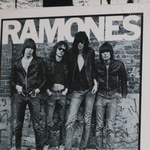 May include: A black and white photo of the Ramones band on a white album cover. The band members are wearing leather jackets and jeans. The album cover has the band's name "RAMONES" printed in large, bold letters.