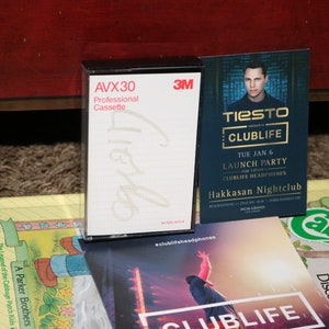 Könnte beinhalten: Ein weißes Kassettenband mit einem schwarzen Etikett, auf dem "AVX30 Professional Cassette" und das 3M-Logo stehen. Das Kassettenband liegt neben einem Flyer für eine Tiesto Clublife Launch Party im Hakkasan Nightclub am 6. Januar. Der Flyer zeigt ein Foto von Tiesto und den Text "Tiesto Presents Clublife".