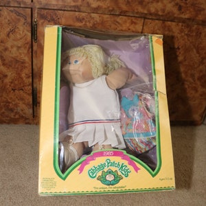 May include: A Cabbage Patch Kids doll in a white tennis dress with a blue and pink striped skirt. The doll has blonde hair and blue eyes. The box is yellow and green with the Cabbage Patch Kids logo and the text "1985" and "I'm unique. I'm adoptable!"  Ages 3 up.