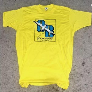 May include: Yellow t-shirt with a blue and white graphic of a seagull flying over the letters "OB" and the text "Ocean Beach California".