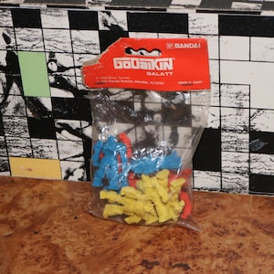 May include: A clear plastic bag containing small, colorful GoDaiKin Galatt action figures. The figures are blue, yellow, and red. The bag has a red and orange cardboard header with the Bandai logo and product information.