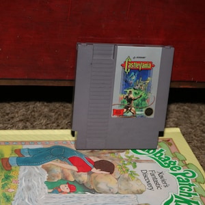 May include: A vintage Nintendo Entertainment System (NES) game cartridge for Castlevania. The cartridge is gray with a black label featuring a colorful illustration of a knight fighting a monster.