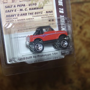 May include: A red and black 1/64 scale die-cast model car of a Suzuki Samurai, hand-built by Prototype. The car is in a clear plastic blister pack with a white background. The packaging includes the text "Hand-built by Prototype 1/64".
