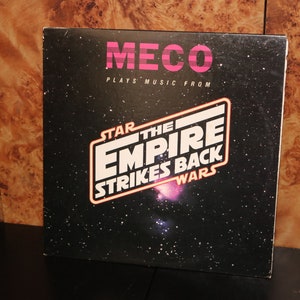 Puede incluir: Una portada de álbum de vinilo negro con un gráfico rosa y blanco del texto "Star Wars: El Imperio Contraataca" con un fondo de galaxia. La portada del álbum también presenta el texto "MECO Plays Music From".