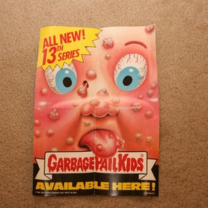 May include: A folded poster advertising Garbage Pail Kids trading cards. The poster features a cartoon character with a red face, large blue eyes, and a long, pink tongue sticking out. The text on the poster reads "All New! 13th Series Garbage Pail Kids Available Here!"
