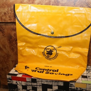 Pode incluir: Bolsa de vinil amarela com uma aba marrom e um fecho de pressão. A bolsa tem um logotipo dos Padres de San Diego com um personagem de desenho animado segurando um taco de beisebol. O texto "Save at Central Savings" está impresso na parte inferior da bolsa.