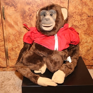 May include: A brown plush monkey wearing a red jacket with a white zipper. The monkey has a smiling face with brown eyes and a brown nose.