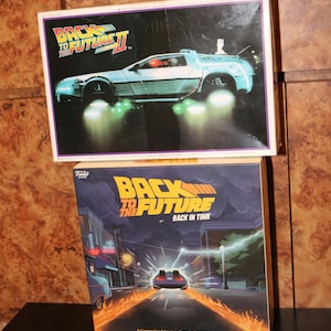 Puede incluir: Dos cajas de juego para el juego de mesa Regreso al futuro. La caja superior presenta una máquina del tiempo DeLorean plateada con luces brillantes. La caja inferior muestra una imagen de dibujos animados de la DeLorean conduciendo por una calle con llamas que salen de las ruedas. El texto en las cajas dice "Regreso al futuro" y "De vuelta en el tiempo".