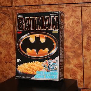 May include: A vintage Batman cereal box with a black background and the Batman logo in gold. The box reads "BATMAN" in large gold letters and includes the text "FREE BATMAN COMIC BOOKS!" The box contains 14 oz (396g) of cereal.