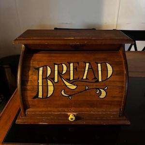 May include: A dark brown wooden bread box with a roll-top door. The front of the door features the word BREAD painted in large, stylized yellow letters with black outlines and decorative flourishes. A small, round yellow knob is centered at the bottom.