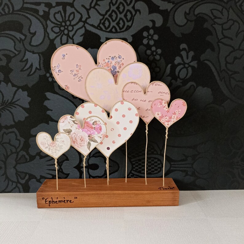 May include: A wooden base with the word "Ephemer" inscribed on it holds a collection of six pink and white paper hearts with floral and polka dot patterns. The hearts are attached to thin wires and arranged in a cascading design.