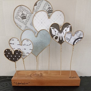 May include: A collection of seven paper hearts in various patterns and colors, including white, gray, and blue, are displayed on a wooden base. The hearts are attached to the base with twine and are arranged in a cluster. The base is labeled "Ephemer" and "Pluche".
