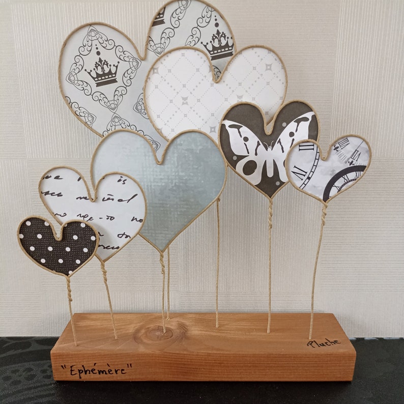May include: A collection of seven paper hearts in various patterns and colors, including white, gray, and blue, are displayed on a wooden base. The hearts are attached to the base with twine and are arranged in a cluster. The base is labeled "Ephemer" and "Pluche".