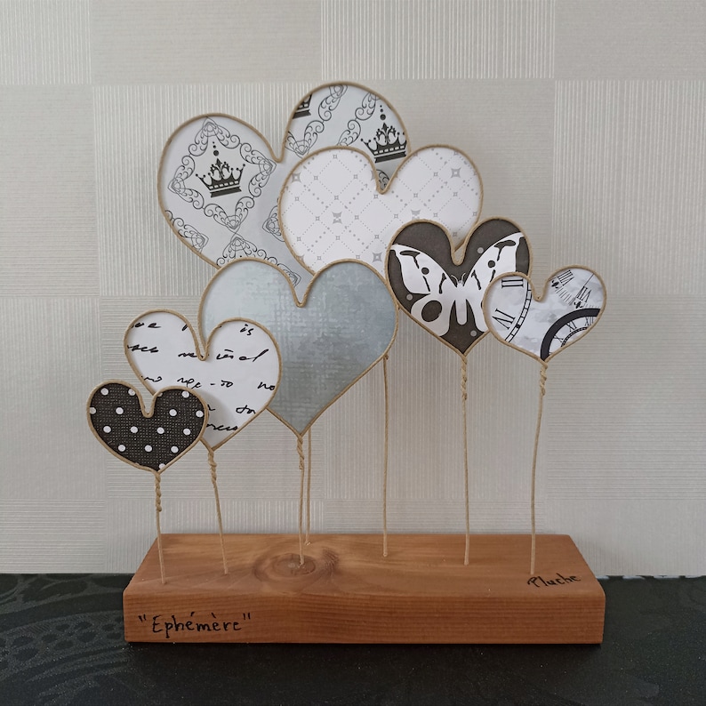 May include: A wooden base with the word "Ephemeral" inscribed on it holds a collection of seven paper hearts. The hearts are decorated with various patterns, including a crown, a butterfly, and a clock face. The hearts are attached to the base with twine.