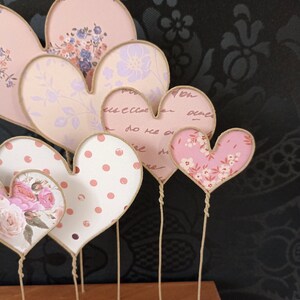 May include: A set of six pink and white heart-shaped decorations with different patterns, each attached to a twine string. The hearts are made of fabric and have a vintage aesthetic.