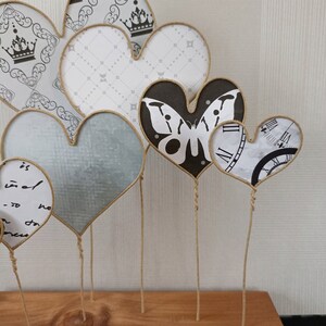 May include: A collection of six decorative paper hearts on wire stems. The hearts are decorated with various patterns, including a butterfly, a clock face, and a geometric design. The hearts are arranged in a cluster on a wooden surface.