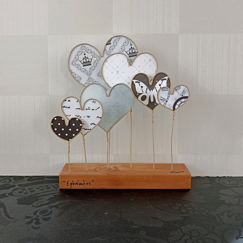 May include: A wooden base with the word "Ephemer" inscribed on it holds a collection of seven paper hearts. The hearts are decorated with various patterns, including polka dots, a butterfly, and a clock face.