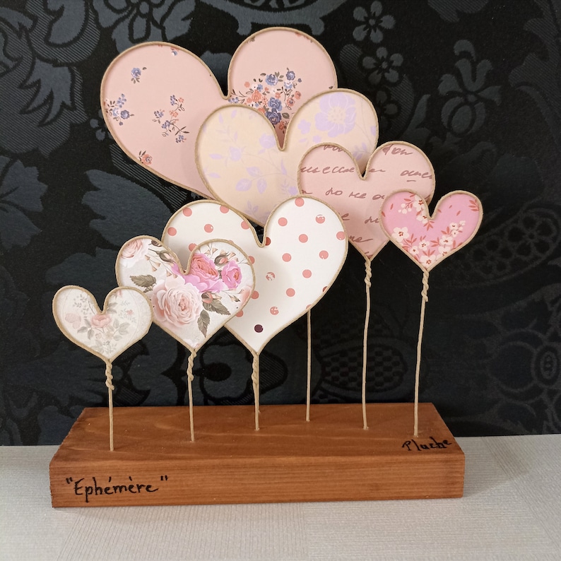 May include: A wooden base with the word "Ephemer" written on it holds a collection of six paper hearts on wire stems. The hearts are decorated with various patterns, including polka dots, floral designs, and handwritten text.